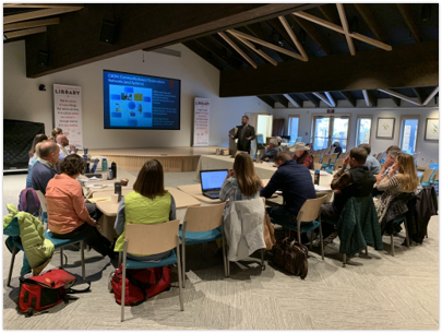 A modern conference room with rows of folding chairs and tables. A large projection screen displays a presentation while numerous attendees listen.
