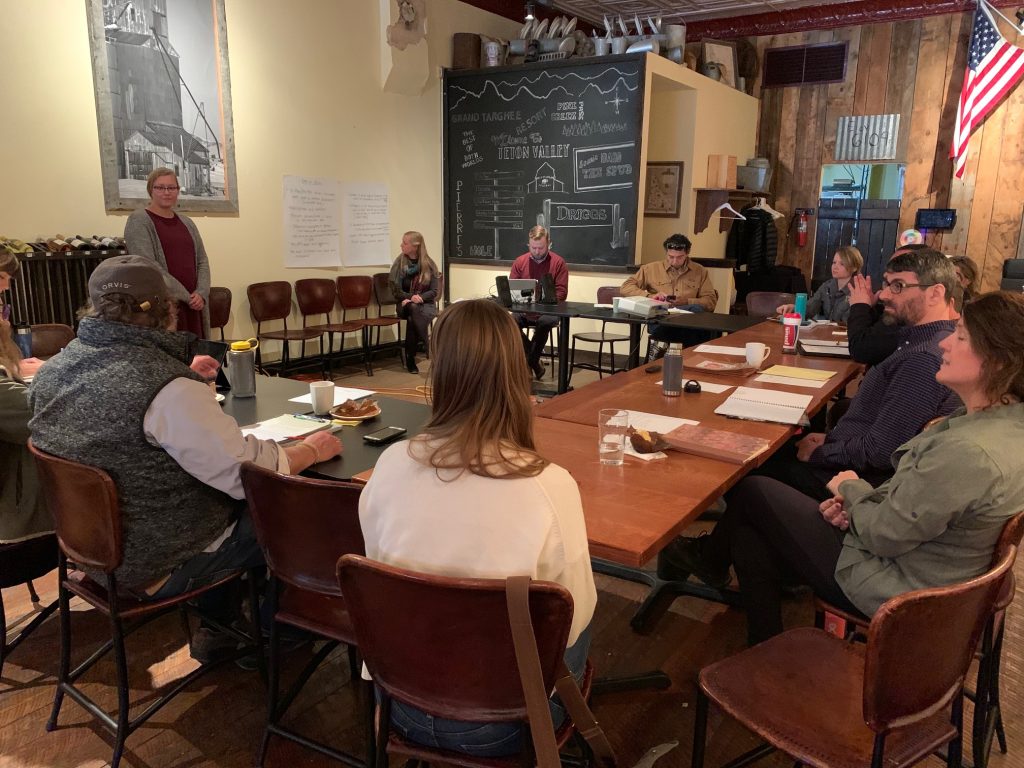 A meeting room with exposed brick walls and vintage decor. A chalkboard displays diagrams and text while attendees sit around tables. A person in a burgundy sweater stands presenting.