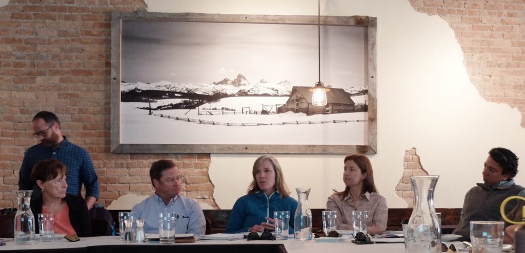 A meeting space with a large panoramic winter landscape photograph. Several people sit at tables with water glasses and notepads.