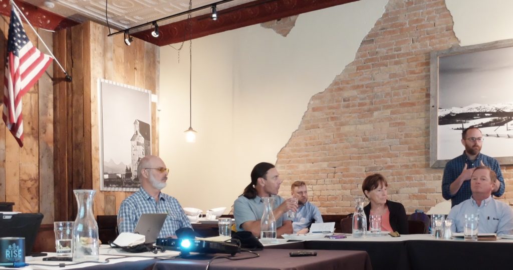 A meeting in progress with attendees seated at tables covered in white tablecloths. Water carafes and glasses are placed throughout. A large winter landscape photograph hangs on the brick wall.